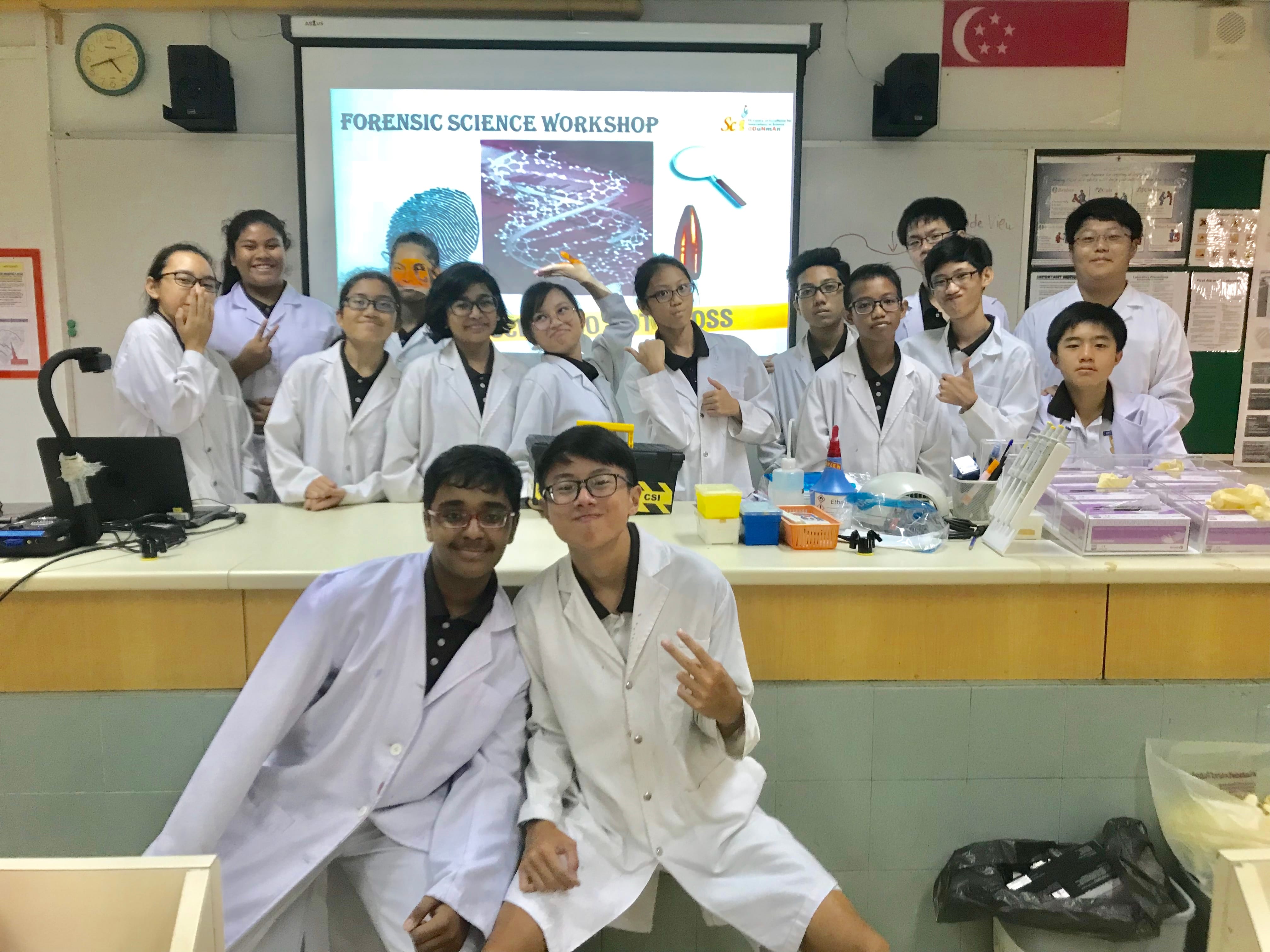 Students in white lab coats pose in a classroom in front of a Forensic Science Workshop projected slide.